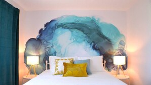 Un dormitorio con un cuadro abstracto grande en la pared, una cama con ropa de cama blanca y dos almohadas decorativas, y lámparas de noche a ambos lados.
