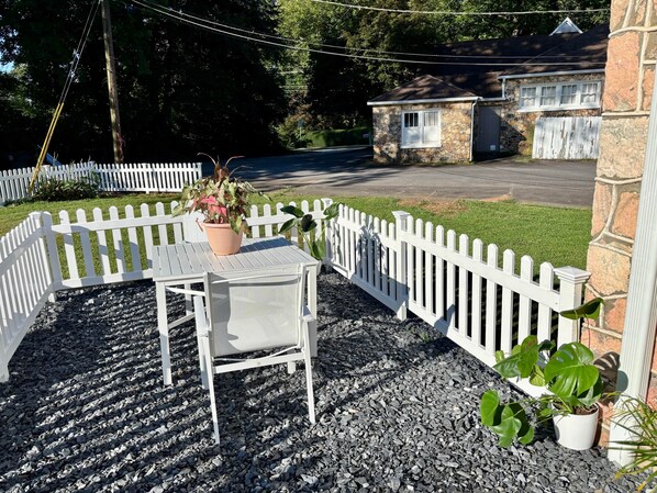 A white outdoor seating area with a table and chairs, surrounded by a white picket fence, and a stone building in the background.