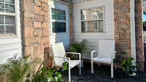 A stone exterior with white window frames and a small patio area with white chairs and potted plants. - Woodward Villa A (Dahlonega)