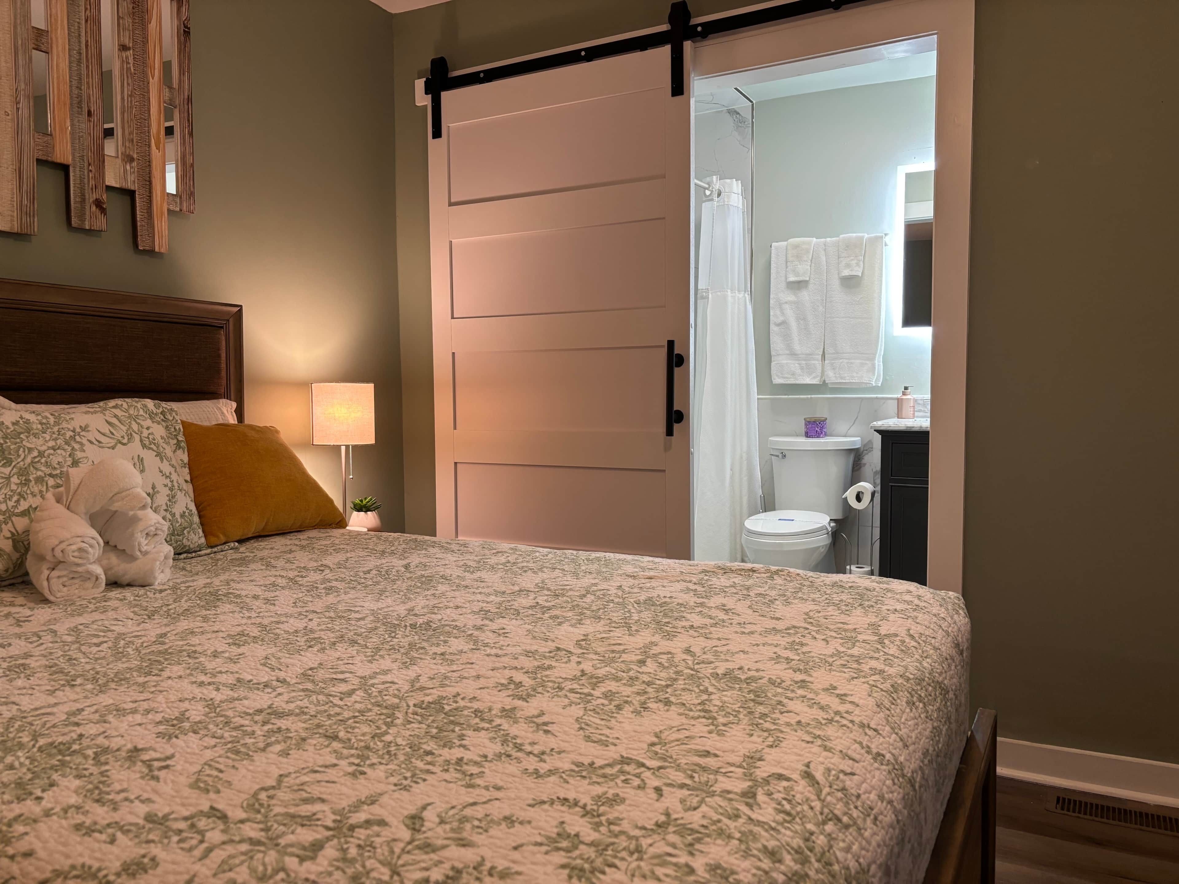 A bedroom with a bed, a nightstand, a lamp, a wooden headboard, a sliding door to a bathroom, and a wall-mounted mirror.