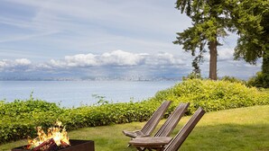 A fire pit with logs, two wooden lounge chairs, and a view of a body of water with a distant city skyline. - Peaceful Beachfront Getaway with Sweeping Water Views (Blaine)