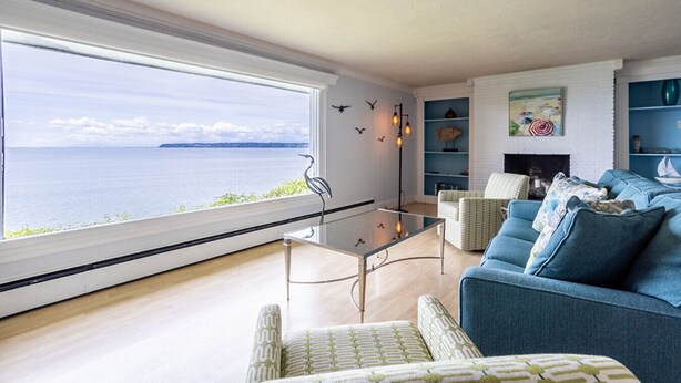 A modern living room with a large window overlooking a body of water, a glass coffee table, a blue sofa, and patterned armchairs. - Peaceful Beachfront Getaway with Sweeping Water Views (Blaine)