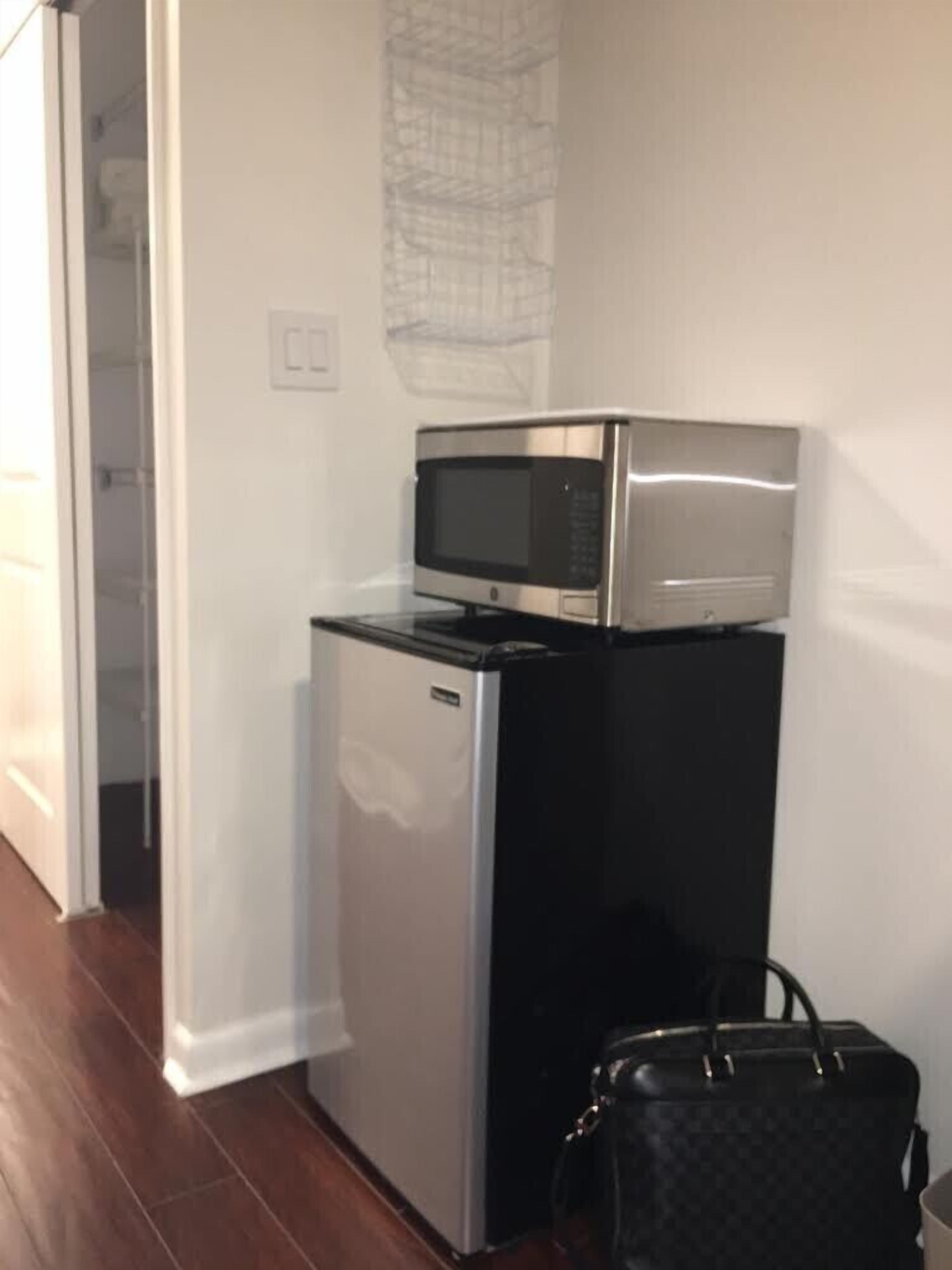 Une petite cuisine avec un micro-ondes posé sur le dessus d’un réfrigérateur, un sac à main noir sur le sol et une porte blanche à l’arrière-plan.