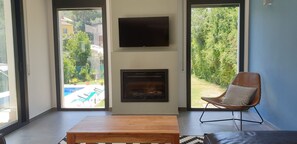 A living room with a fireplace, a TV mounted on the wall, and a view of a pool and greenery outside. - Maison Moderne Piscine Privée 4kms des Plages (Calafell)
