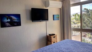 A bedroom with a bed, a flat-screen TV mounted on the wall, a small wooden side table, and a window with a view of palm trees. - AMAZING HOTEL ZONED BEACH FRONT CONDO ON SUGAR BEACH.  RESERVE WITH CONFIDENCE! (Kihei)