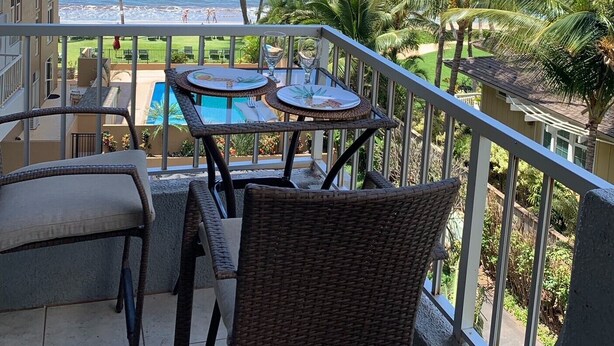 A balcony with wicker furniture overlooking a pool and ocean view, with palm trees and a clear sky. - AMAZING HOTEL ZONED BEACH FRONT CONDO ON SUGAR BEACH.  RESERVE WITH CONFIDENCE! (Kihei)