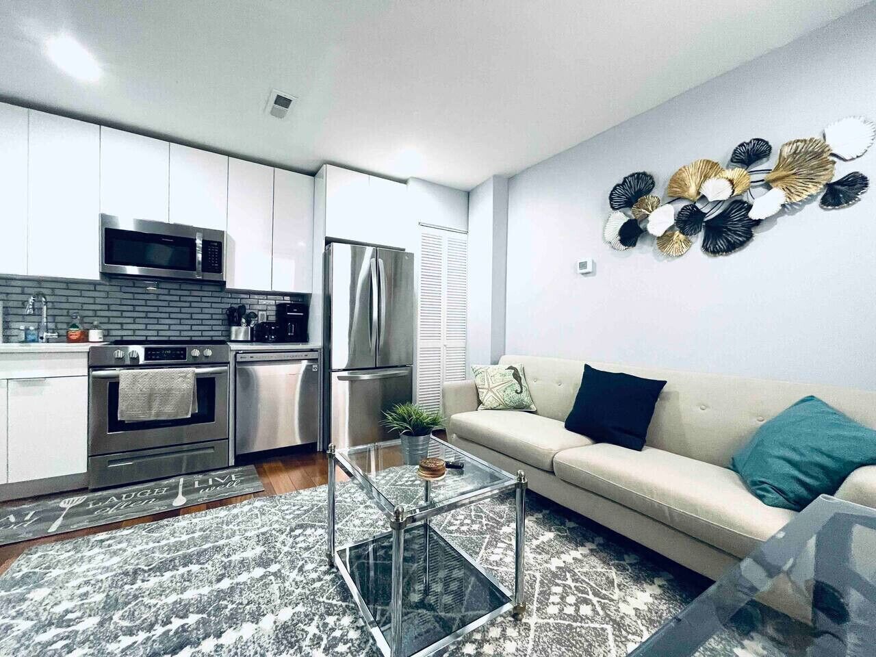 Una cocina moderna con electrodomésticos de acero inoxidable, una mesa de centro de vidrio y un sofá color beige con cojines verde azulado.
