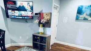 Una sala moderna con televisor de pantalla plana, una mesa de centro de vidrio, un tapete estampado, piso de madera y un comedor con un mantel individual.