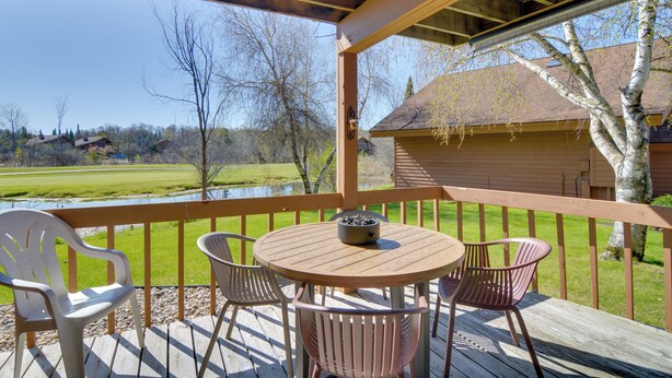 A wooden deck with a round table and four chairs, overlooking a grassy area and a body of water. - Schuss Mountain Resort Condo: Steps to Golf! (Mancelona)