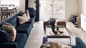 Un salon moderne avec un canapé bleu, une table basse agrémentée d’objets décoratifs et une vue sur un balcon aménagé avec du mobilier d’extérieur.