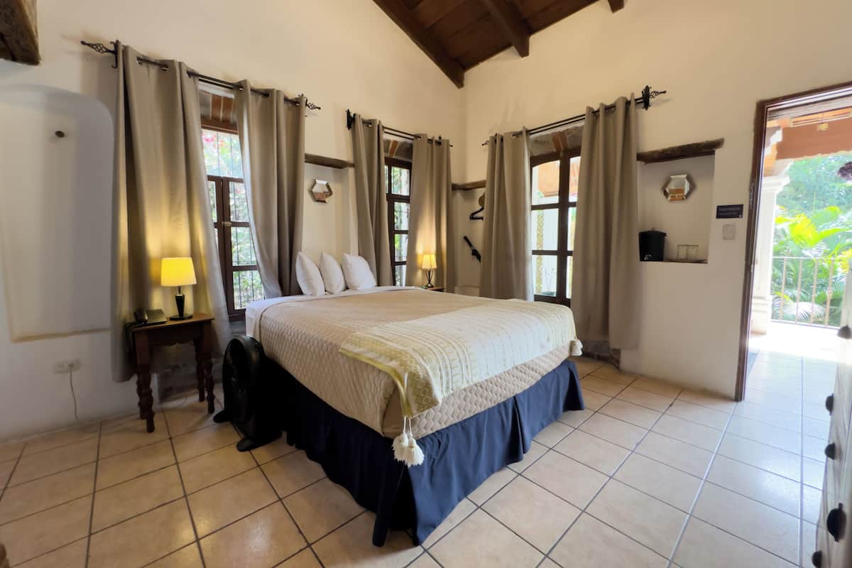 Une chambre à coucher avec un grand lit, des rideaux, une table de chevet avec une lampe et une vue sur la verdure à l’extérieur.