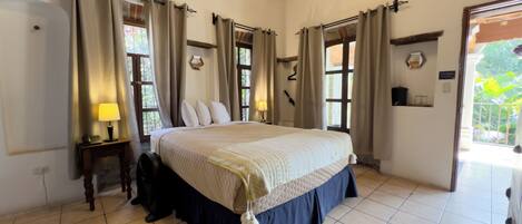 A bedroom with a large bed, curtains, a bedside table with a lamp, and a view of greenery outside.