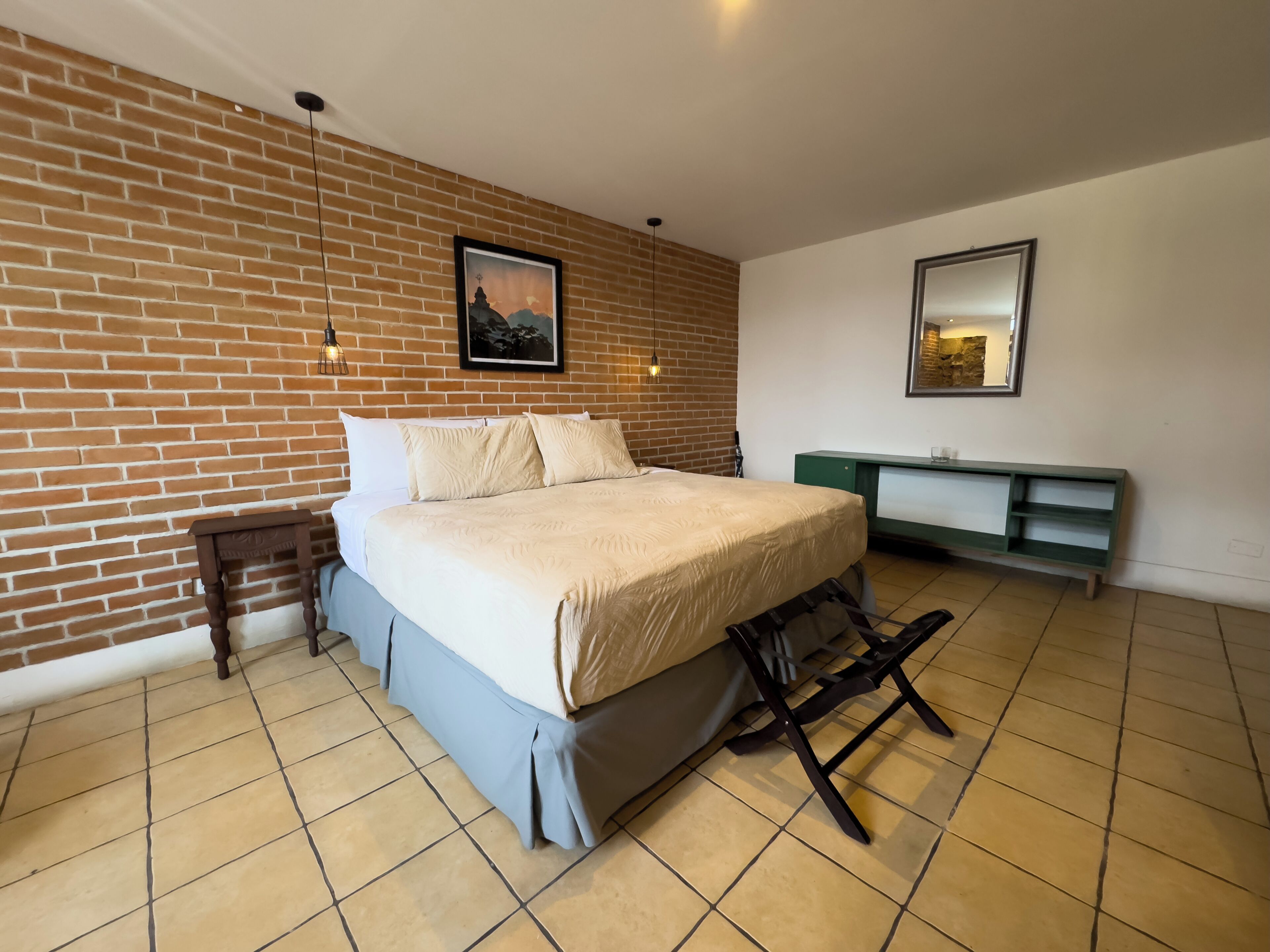 Un dormitorio con pared de ladrillo, una cama con cubrecama blanco, un banco verde y un cuadro de un paisaje.