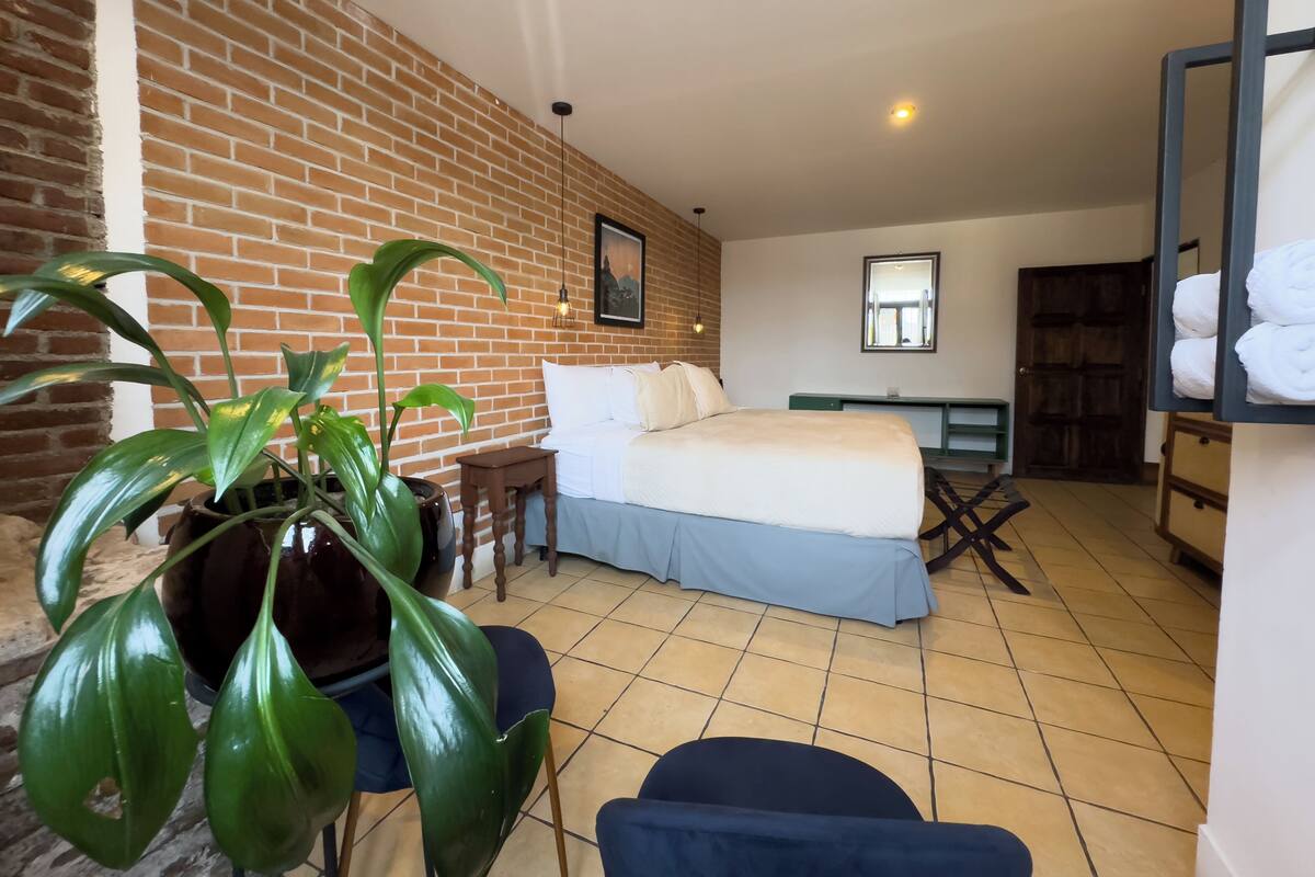 Une chambre d’hôtel avec un mur en briques, un lit, un bureau, une chaise, une plante en pot et un miroir.