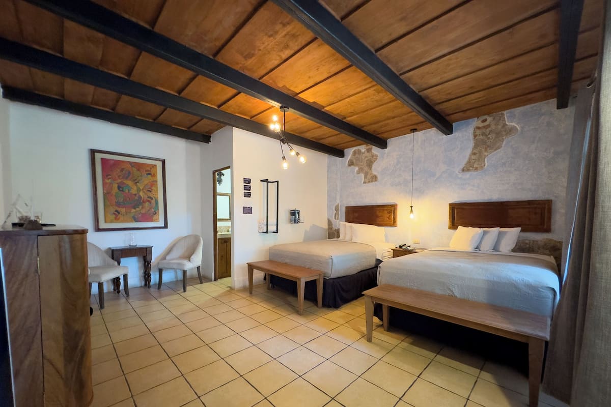 Une chambre d’hôtel avec deux lits, un plafond en bois, un sol carrelé, un bureau avec une chaise et un tableau au mur.