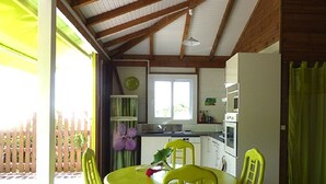 A covered patio with a round yellow table and matching chairs, a kitchen area with white cabinets and a window, and a wooden fence in the background.