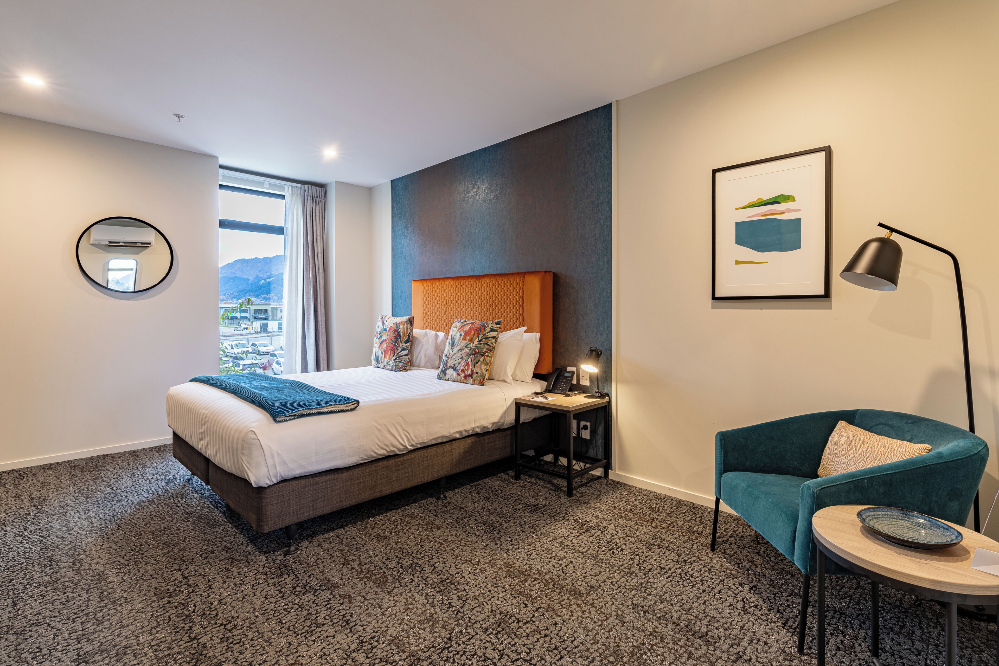 A hotel room with a bed, a desk, a chair, a lamp, a framed picture, and a view of the outside.