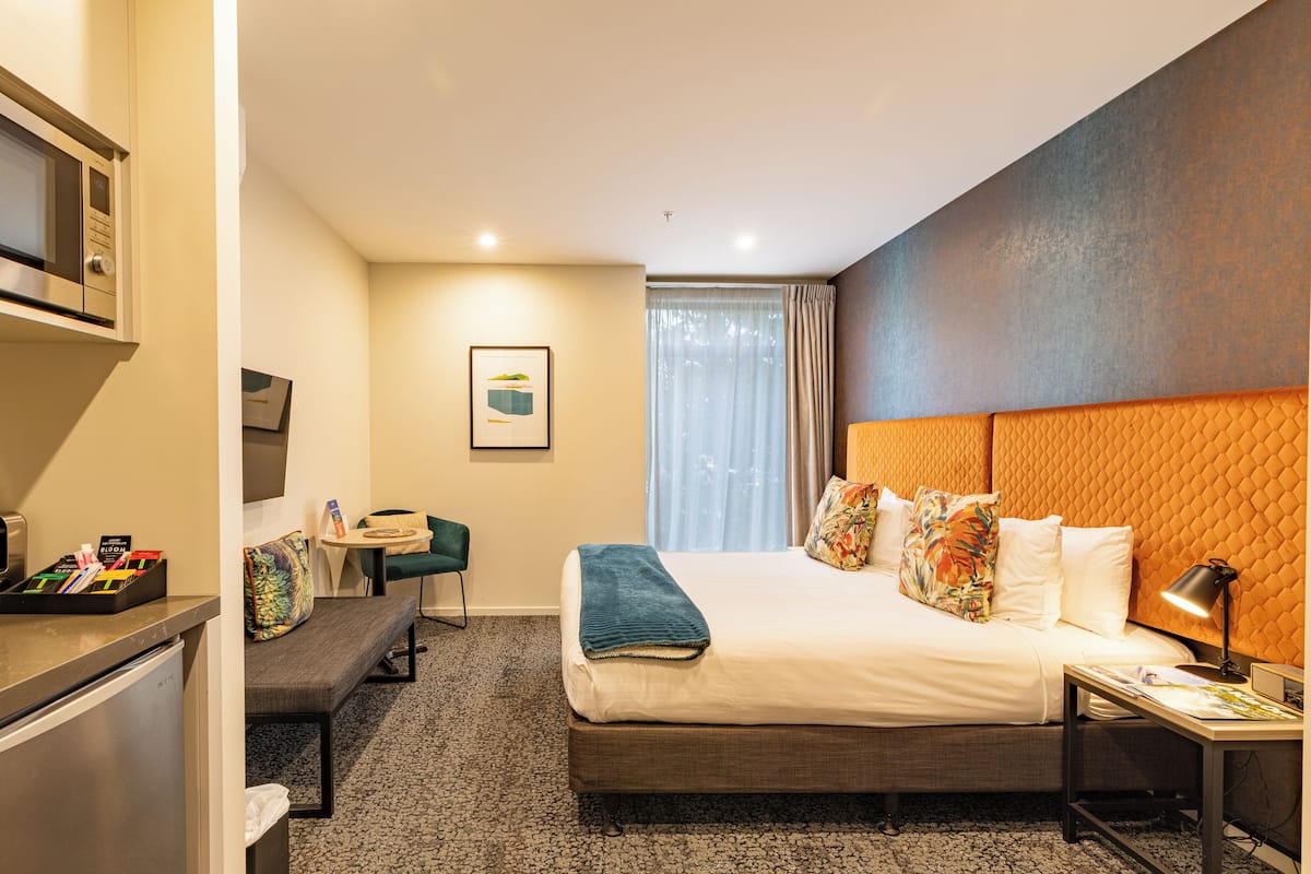 A hotel room with a bed, bedside table, desk, chair, and a kitchenette area.