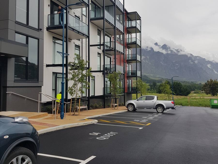 A multi-story building with balconies, a parking area, and a mountainous backdrop.