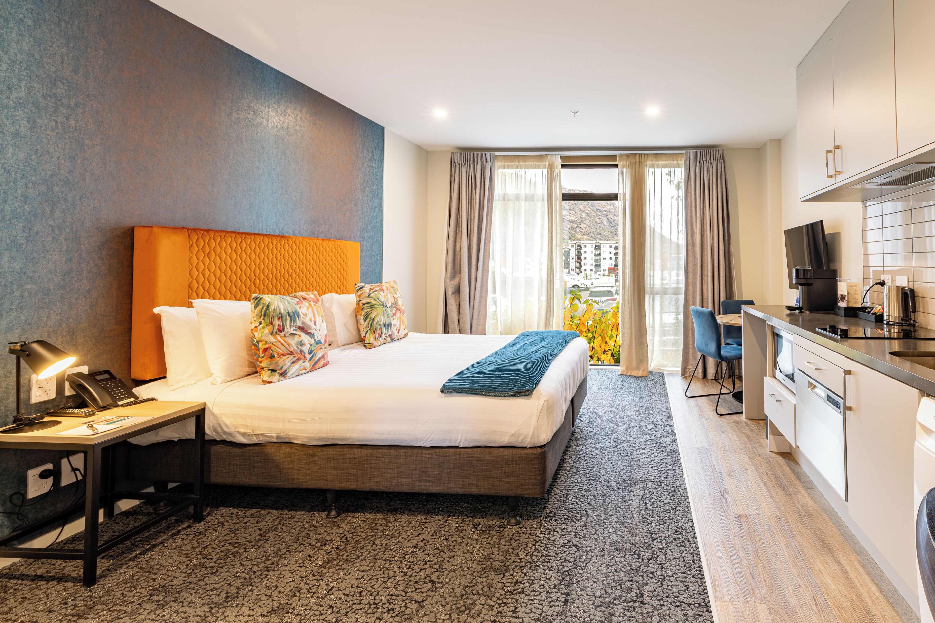 A hotel room with a bed, bedside table with a lamp, a desk with a computer, a kitchenette, and a view of the outside.