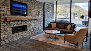 Una sala de estar moderna con una pared de piedra, una chimenea, un televisor, un sofá, una mesa de centro y vistas a una montaña y edificios a través de amplios ventanales.