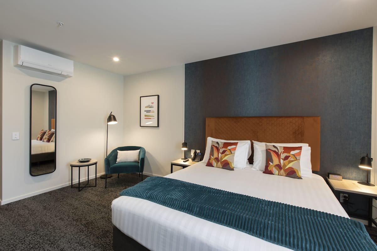 A hotel room with a large bed, a desk, a chair, a mirror, and an air conditioning unit.