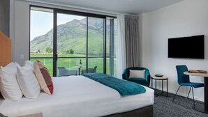Habitación de hotel con una cama grande, televisión, balcón con vistas a montañas y zona de comedor con sillas y mesa.