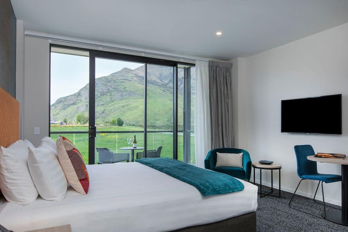 A hotel room with a large bed, a TV, a balcony with a view of mountains, and a dining area with chairs and a table.