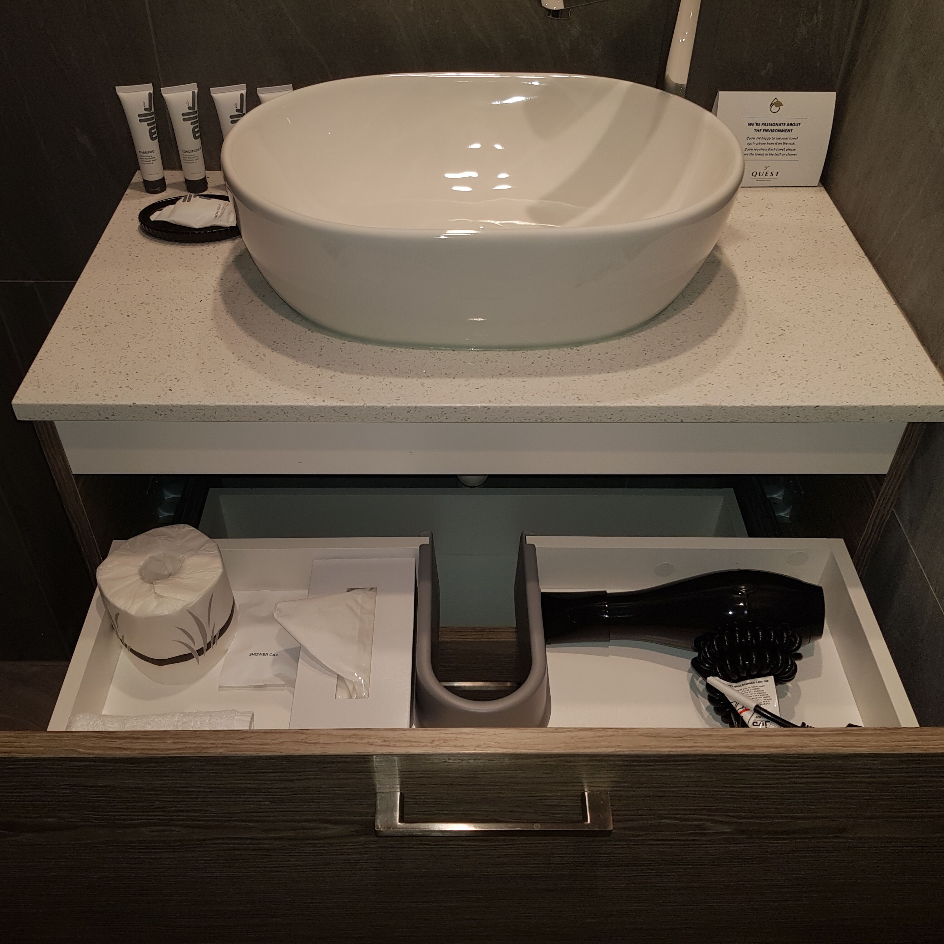 A bathroom with a large white sink, toiletries, and a shower cap in a drawer.