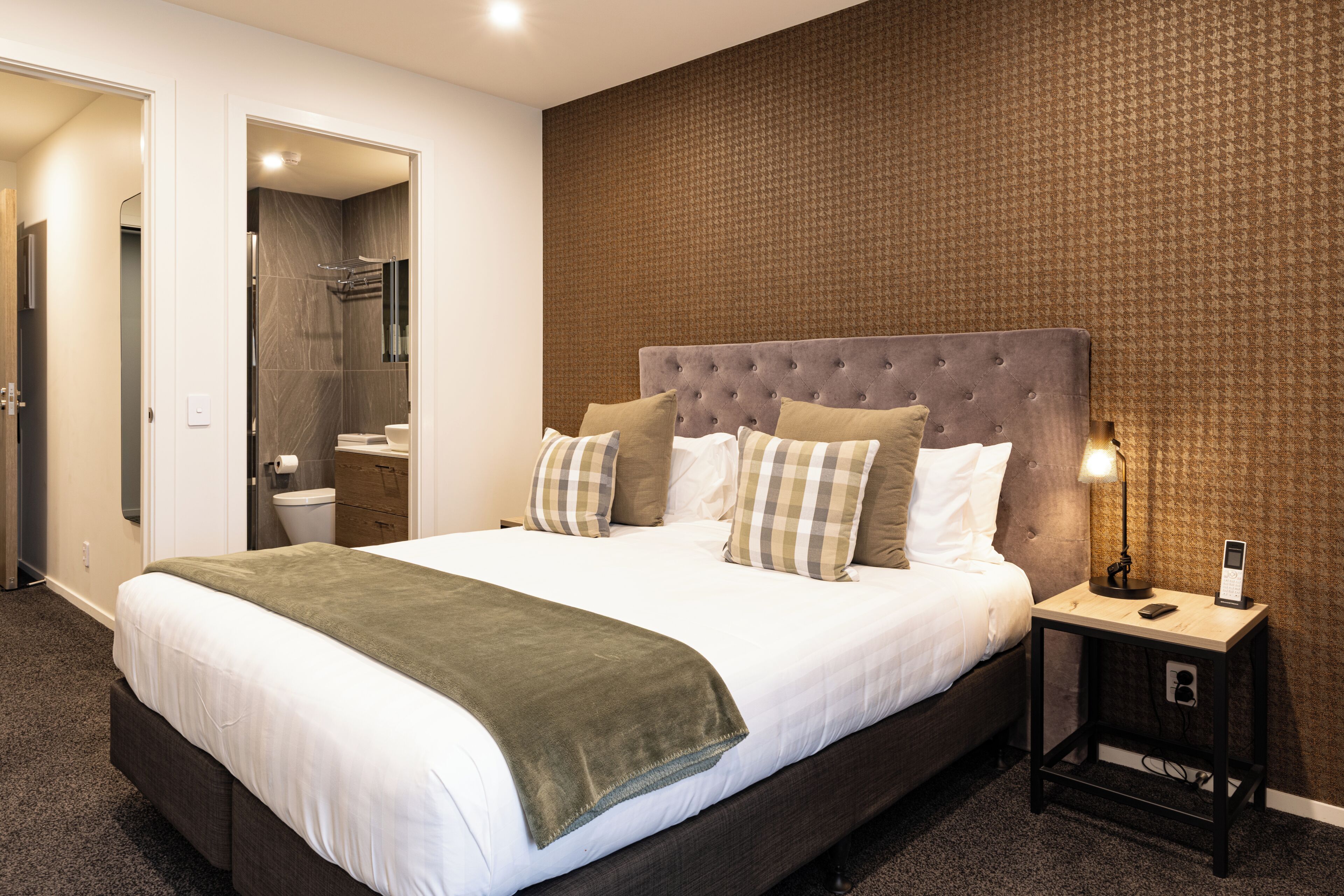 A modern hotel room with a large bed, a bedside table with a lamp, and a bathroom visible through an open door.