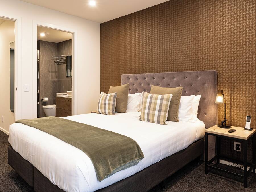 A modern hotel room with a large bed, a bedside table with a lamp, and a bathroom visible through an open door.