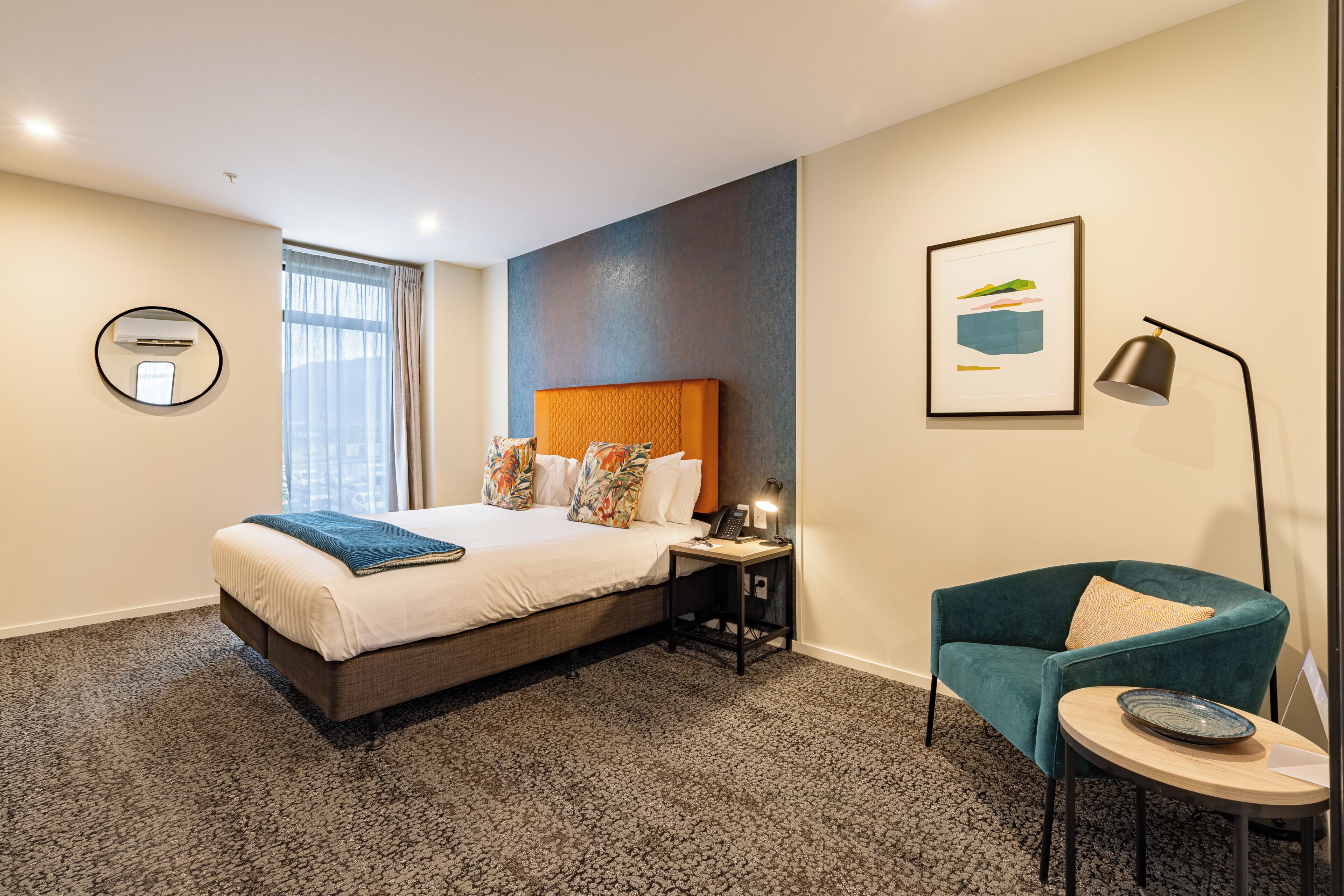 A hotel room with a bed, a desk, a chair, a lamp, and a framed picture on the wall.