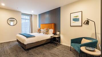 A hotel room with a bed, a desk, a chair, a lamp, and a framed picture on the wall.