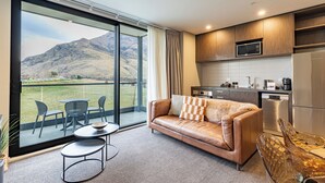 Una sala moderna con sofá, mesa de comedor y sillas, una cocina equipada y un gran ventanal corredera que da a un balcón con vistas a la montaña.