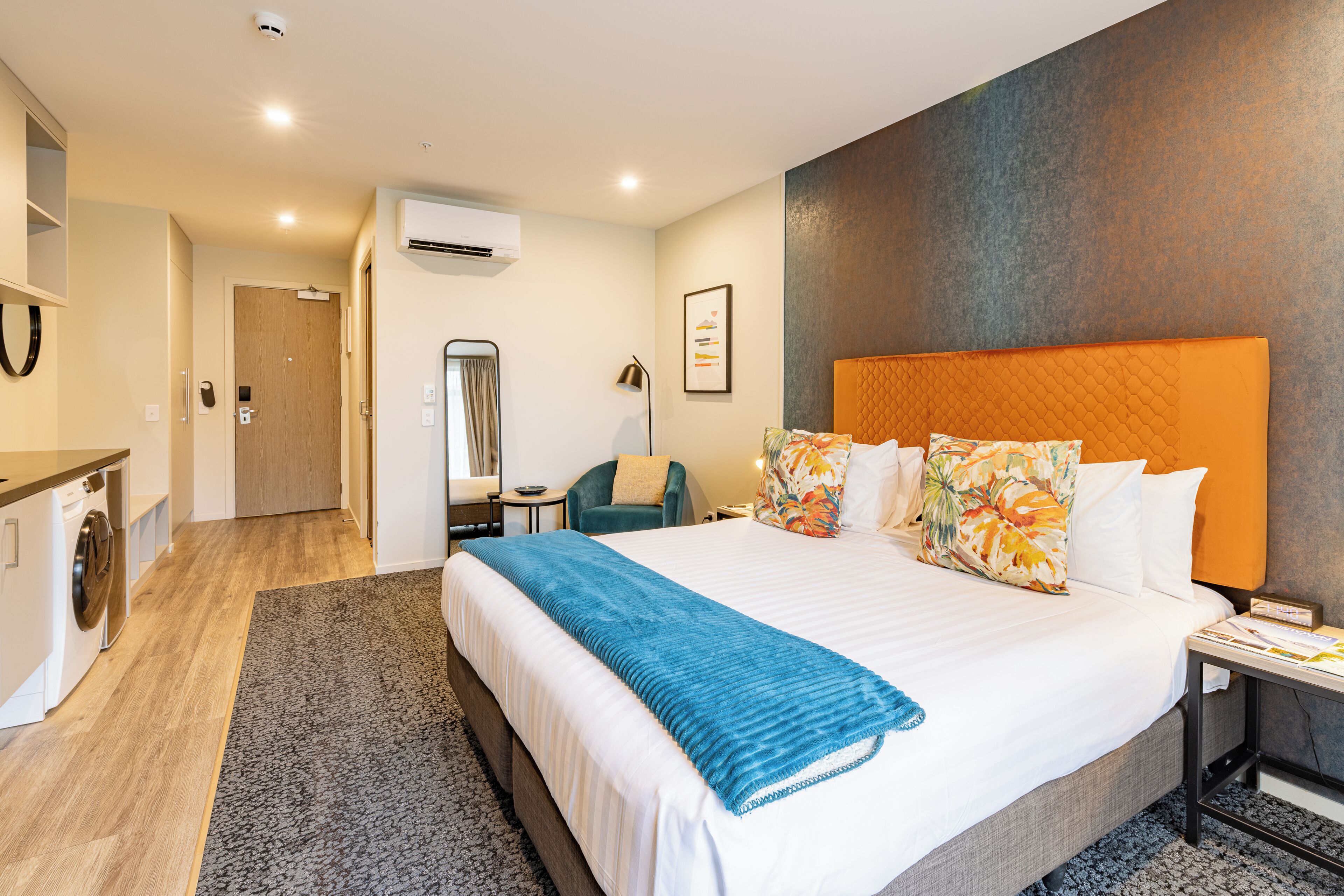 A hotel room with a large bed, a desk, a chair, an air conditioning unit, and a laundry area.