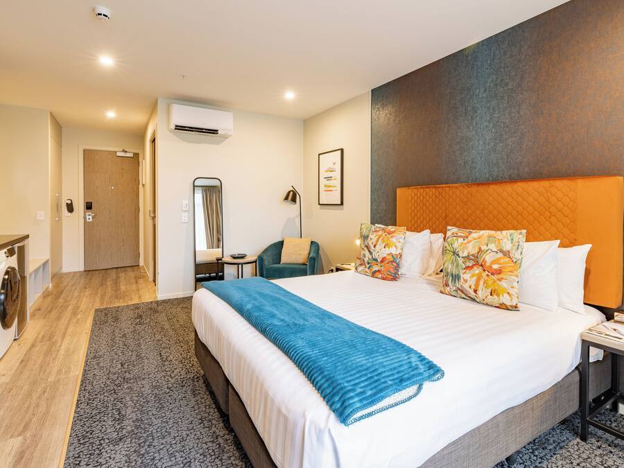 A hotel room with a large bed, a desk, a chair, an air conditioning unit, and a laundry area.