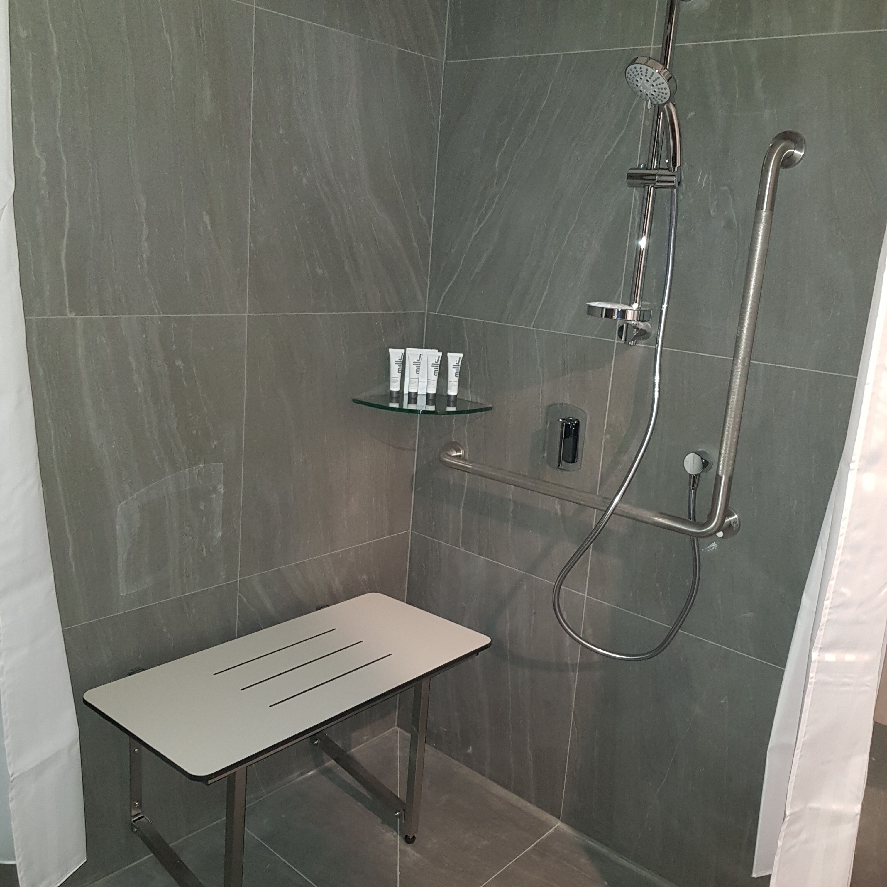 A shower area with a glass shelf holding toiletries, a handheld showerhead, and a handheld shower hose.