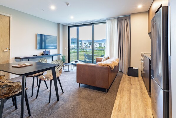 A modern hotel room with a dining area, a sofa, a flat-screen TV, and a large window offering a view of the building's exterior.