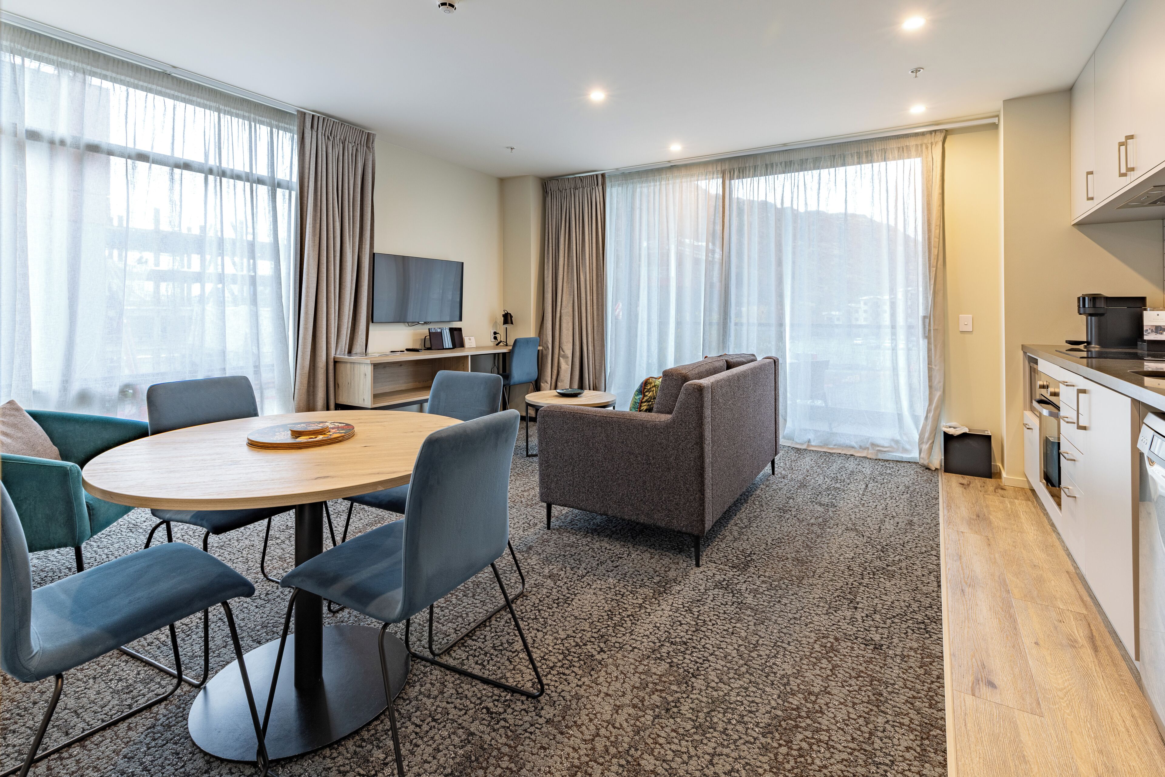 A modern hotel room with a dining area, a sofa, a TV, and a kitchenette.