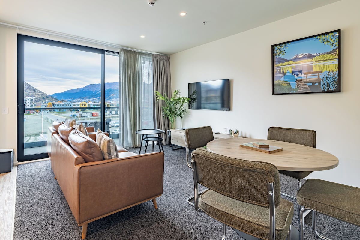 A modern hotel room with a large window, a sofa, a dining table, and a painting.