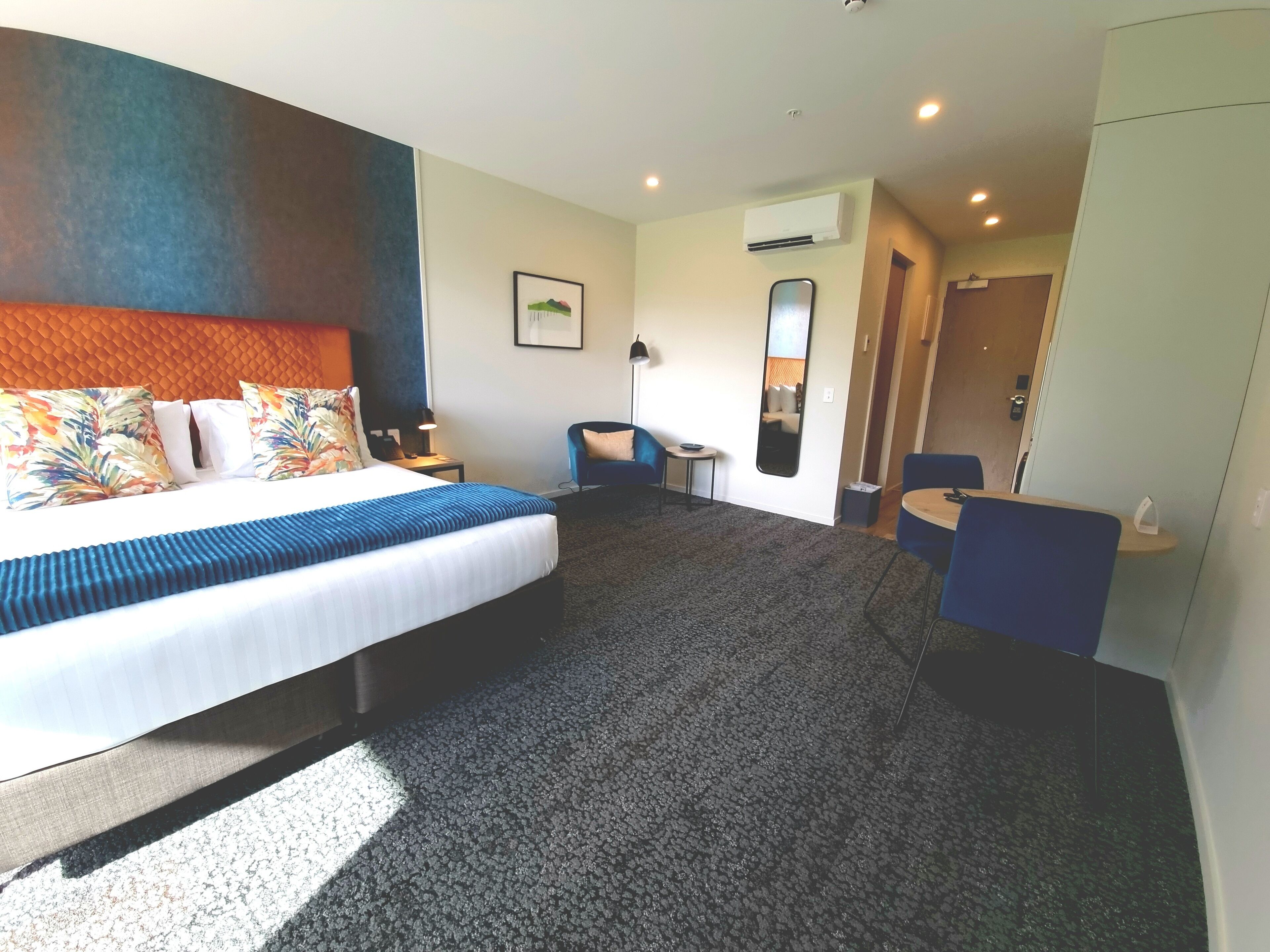 A hotel room with a bed, a desk, a chair, a small table, an air conditioning unit, and a mirror.