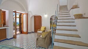 A spacious room with a tiled floor, a wicker chair, a wooden door, and a staircase with a glass railing. - Sweet Home (Praiano)