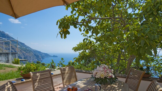 A table set outdoors with a view of the sea, surrounded by wooden chairs, a flower arrangement, and a tray with food and drinks. - Sweet Home (Praiano)