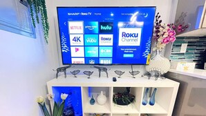Televisão de tela plana presa em uma parede branca, exibindo diversos serviços de streaming.