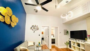 Uma sala de estar moderna com um sofá, uma mesa de centro e um ventilador de teto. Há uma televisão à direita e uma estante de livros abaixo.