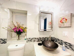 A bathroom with a white toilet, a vessel sink with a black basin, a mirror above the sink, and a floral arrangement on the left. - Downtown City Hall near Kimmel Center 2BD Suite (Philadelphia)