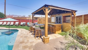 Area piscina con un bar in legno e posti a sedere, circondata da un giardino recintato con lettini.