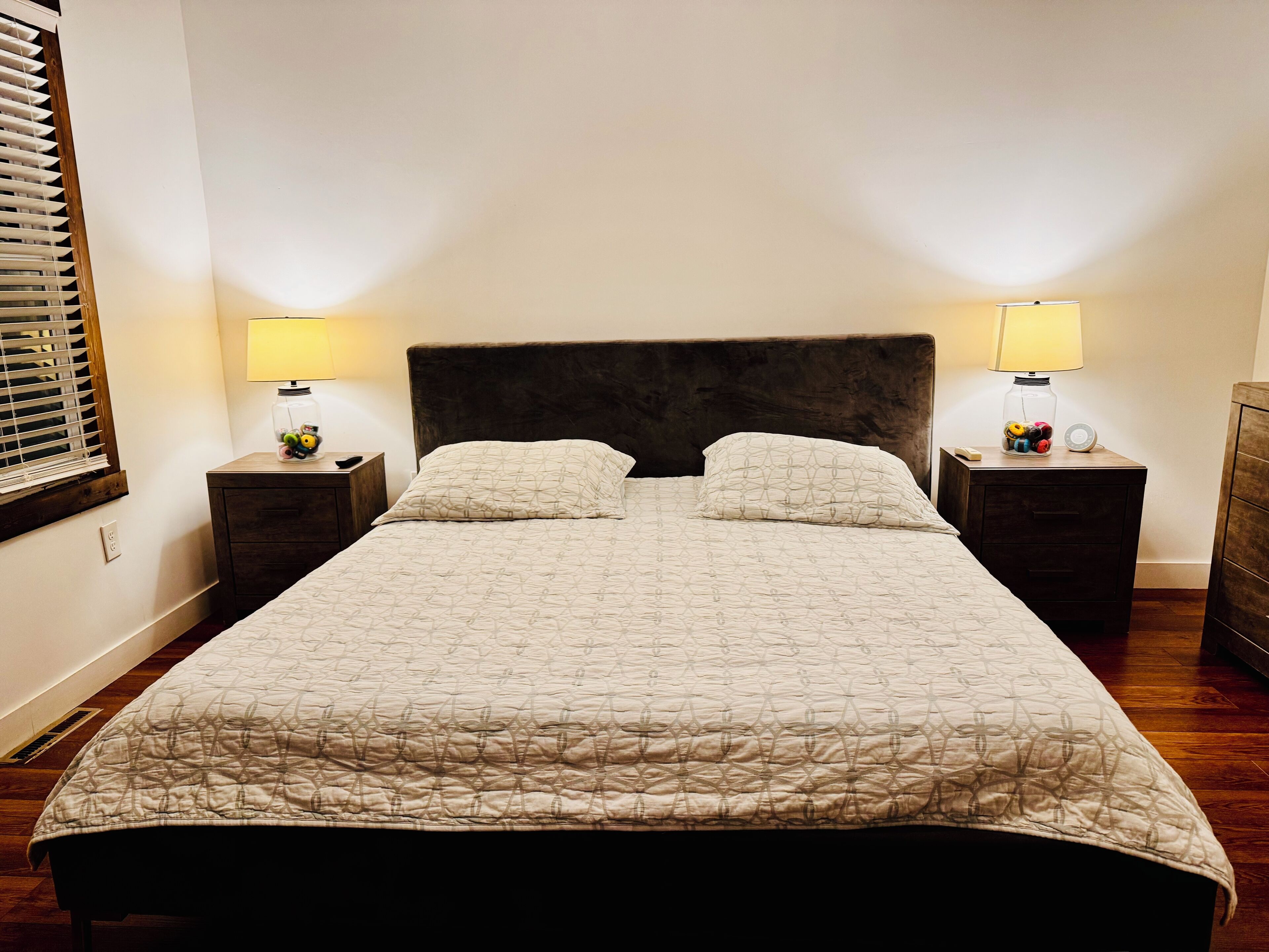 Una cama bien tendida con una colcha estampada y dos almohadas, flanqueada por dos mesitas de noche con lámparas y objetos decorativos.
