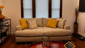 Ein Wohnzimmer mit einem beigefarbenen Sofa, gelben Kissen, einem Holztisch und einem Fenster mit Jalousien.