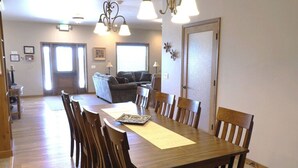 A dining area with a wooden table and chairs, a kitchen in the background, and a living room with a couch and lamps. - Greystone Crossing Suite #11 (Heyburn)